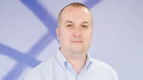 Mind Cymru Simon Jones, a white man with shaved brown/grey hair, looks into the camera with a slight smile. He is wearing a light blue shirt and standing in front of a white and blue background.