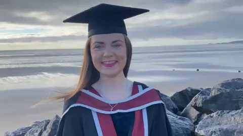 Francesca Murphy Francesca Murphy in graduation gown and cap, standing and looking at the camera with Swansea beach behind her. She smiles.