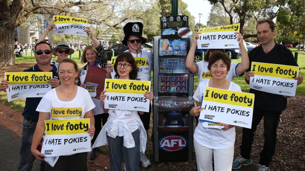 Protesters holding signs reading "I love footy. I hate pokies" stand near a replica poker machine in the park outside of the Melbourne Cricket Ground in Melbourne, Australia.